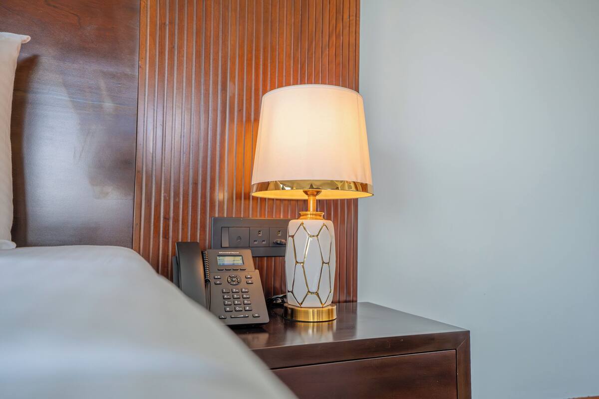 A hotel room with a bed, a bedside table with a lamp and a phone, and a wooden panel wall.