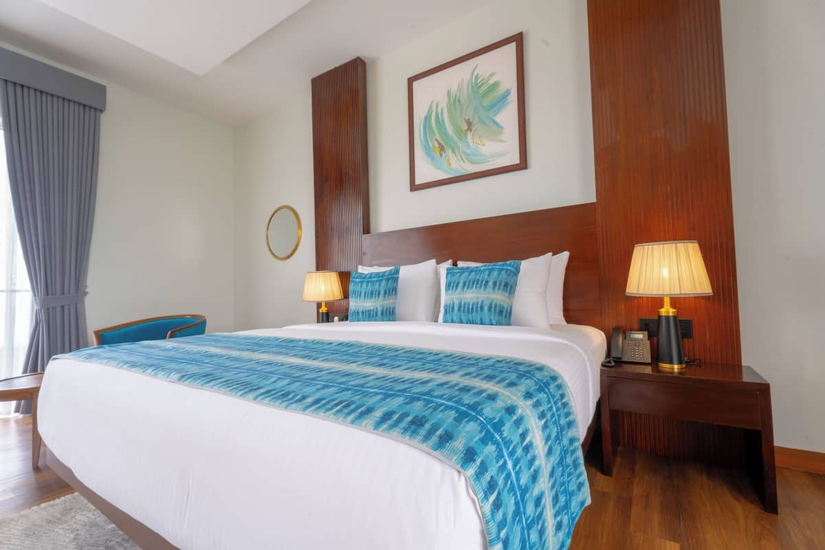 A hotel room with a large bed, two bedside tables with lamps, a wooden headboard, a framed artwork on the wall, and a window with curtains.