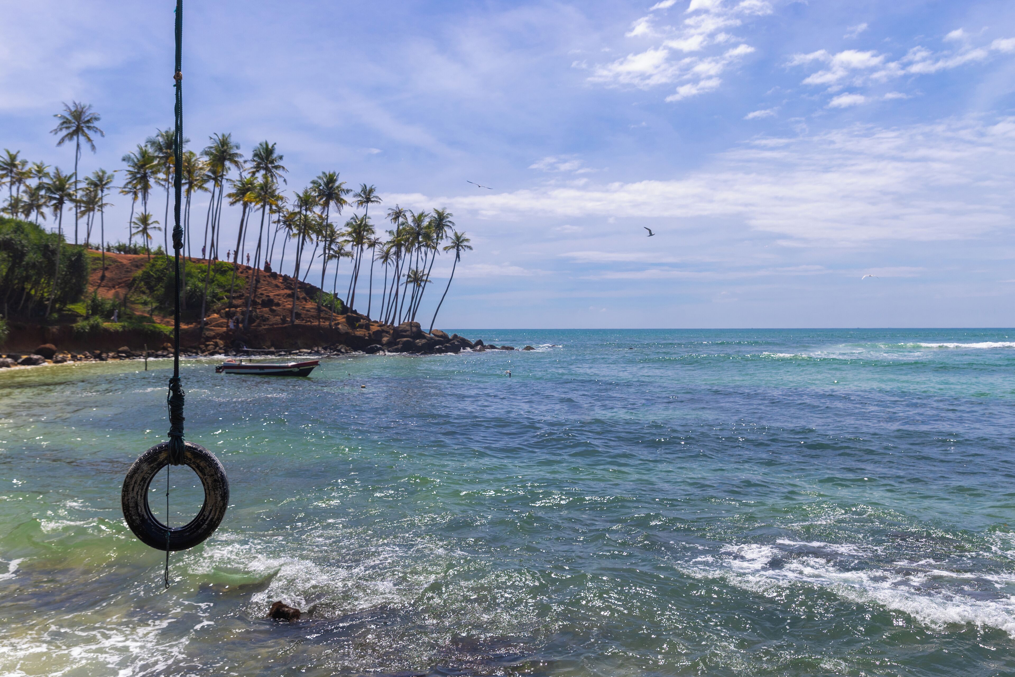 Una playa con aguas turquesas cristalinas, palmeras y un barco a lo lejos.