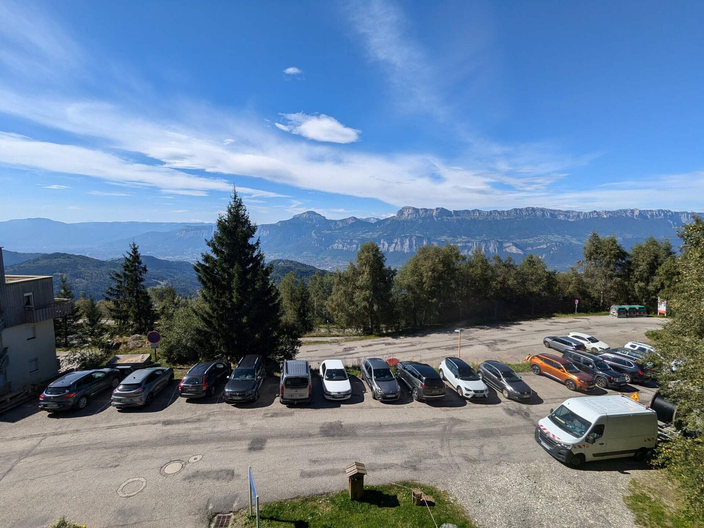 A parking lot with multiple cars and a van, surrounded by trees and mountains in the background.