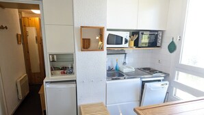 Une cuisine compacte avec des meubles blancs, un four à micro-ondes, une cuisinière et un évier. On y trouve un petit tabouret en bois et une table.