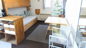 Une pièce compacte et bien aménagée, comprenant un lit, un bureau et une chaise. On y trouve une fenêtre avec des rideaux, un radiateur et une horloge au mur.