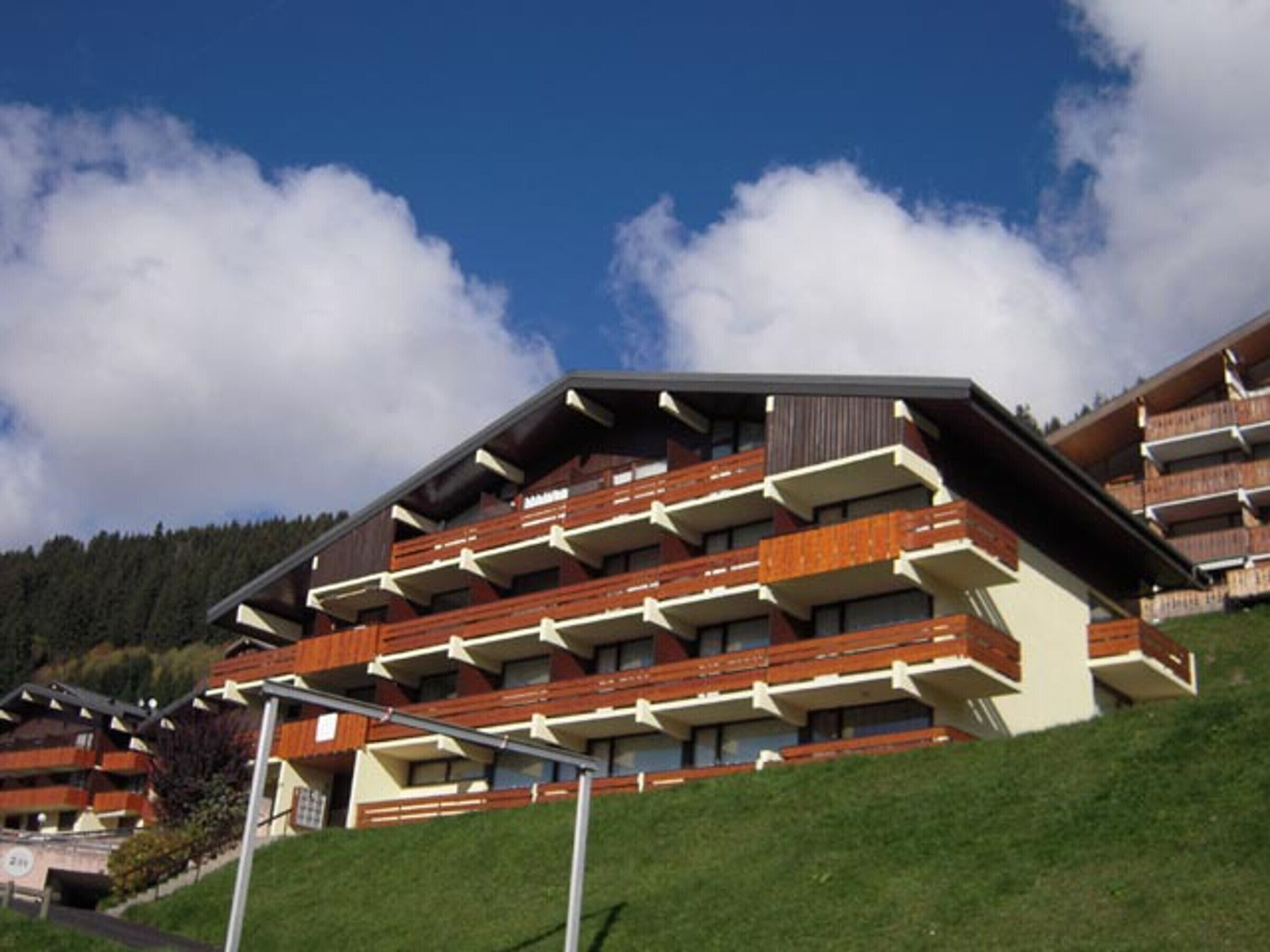 Un hotel de varios pisos con balcones, con un cielo despejado y una colina boscosa de fondo.