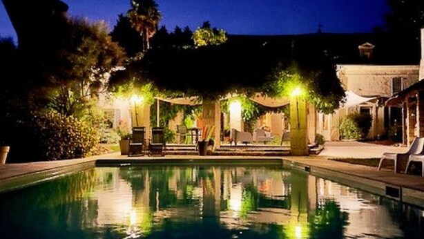 Un espace piscine doté de chaises longues et d’une pergola, entouré de végétation et d’un bâtiment.