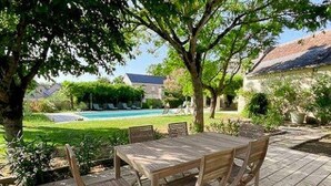 Une terrasse extérieure en bois, avec une table et des chaises, entourée de verdure et avec une piscine en arrière-plan.