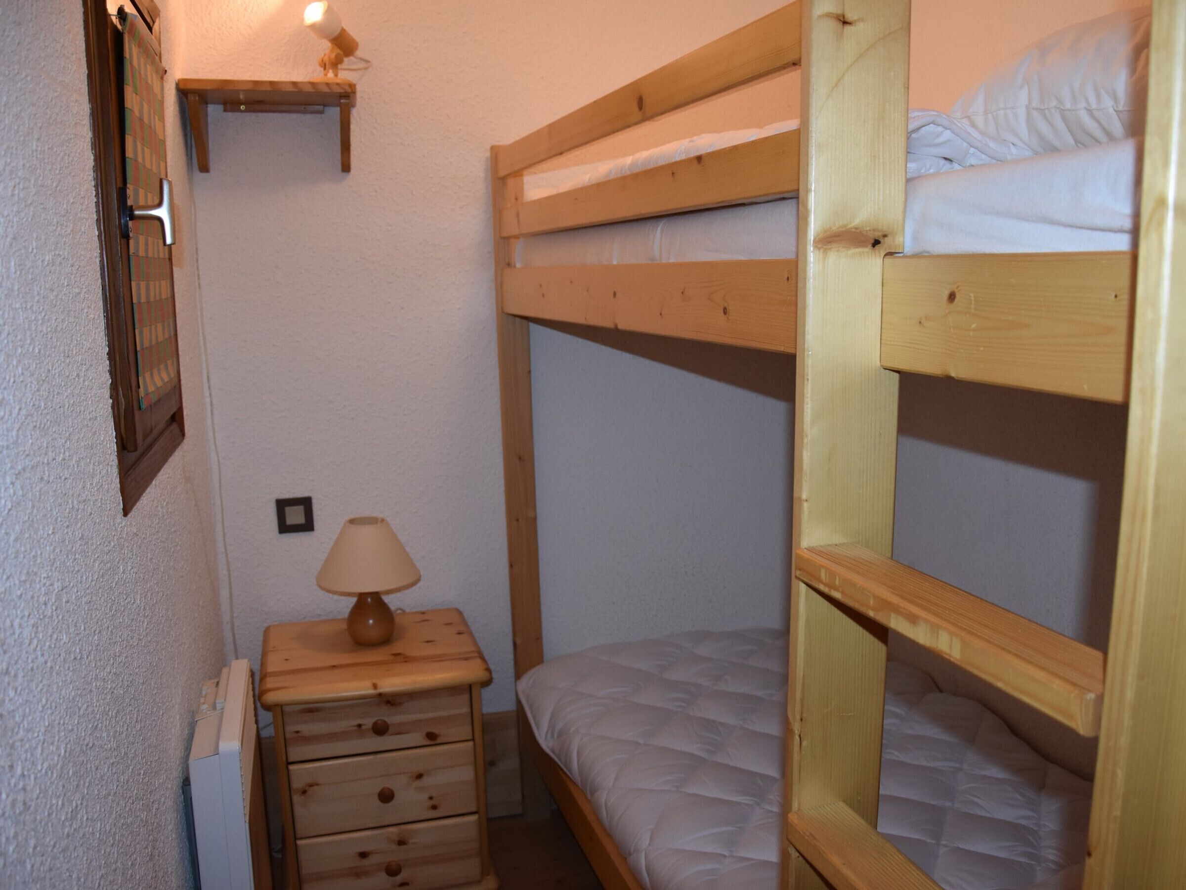 Une chambre avec un lit superposé, une commode en bois, une lampe et une petite étagère.