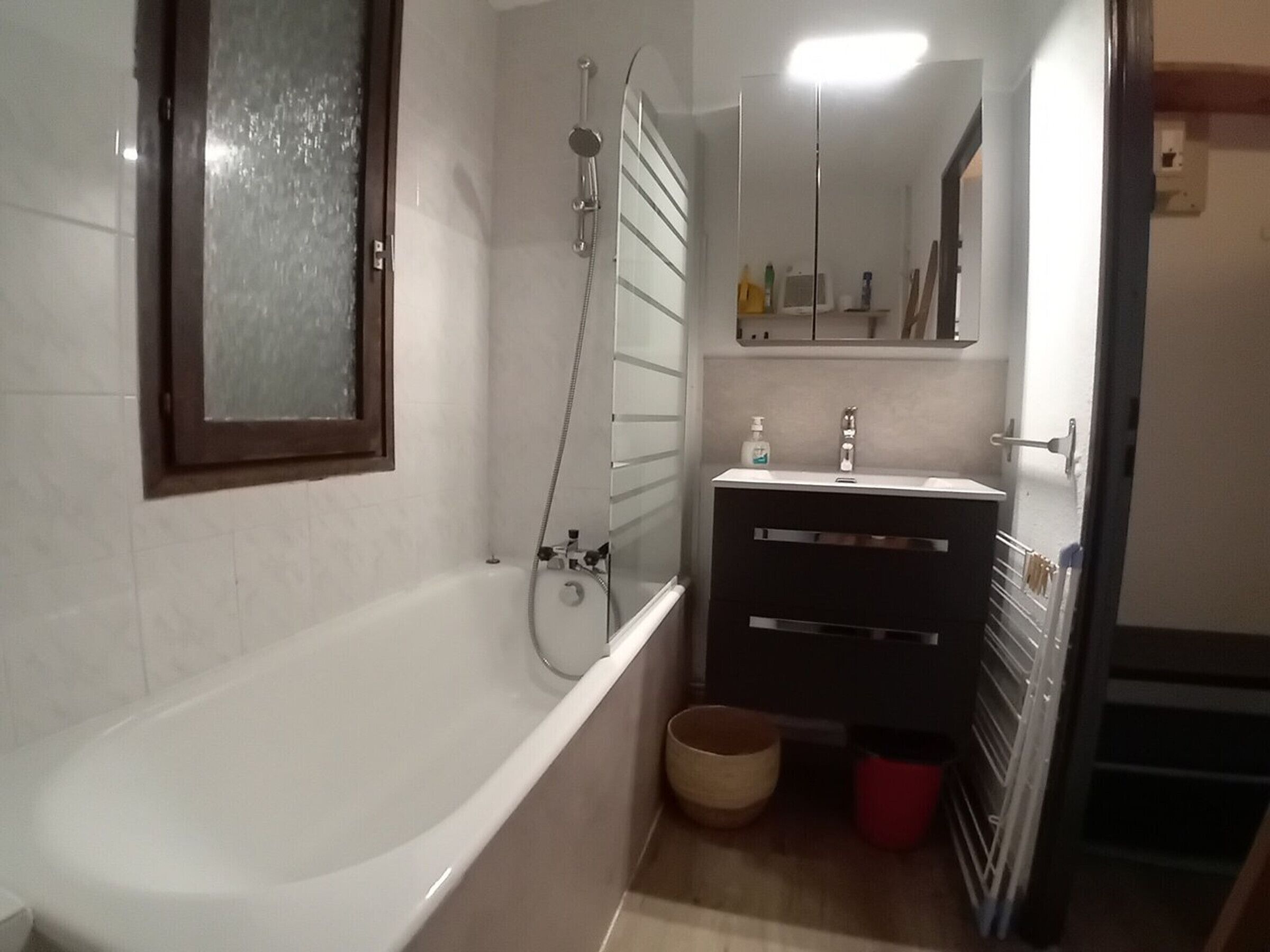Un baño con bañera, ducha, lavamanos y espejo sobre él, además de un taburete de madera.