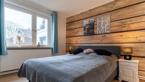 A bedroom with a bed, a nightstand, a window with curtains, and a wooden wall with a picture. - Ferienwohnung in Busum (Büsum)