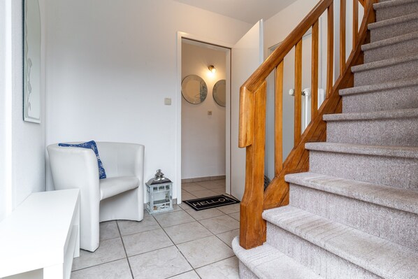 A staircase with wooden railings leading to an upper floor. A white chair with a blue cushion is positioned near a white table. A small, clear lantern is placed on the floor. - Ferienwohnung in Busum (Büsum)