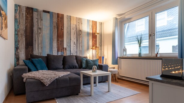A modern living room with a grey sectional sofa, a white coffee table, and a large window with a view of the outside. - Ferienwohnung in Busum (Büsum)