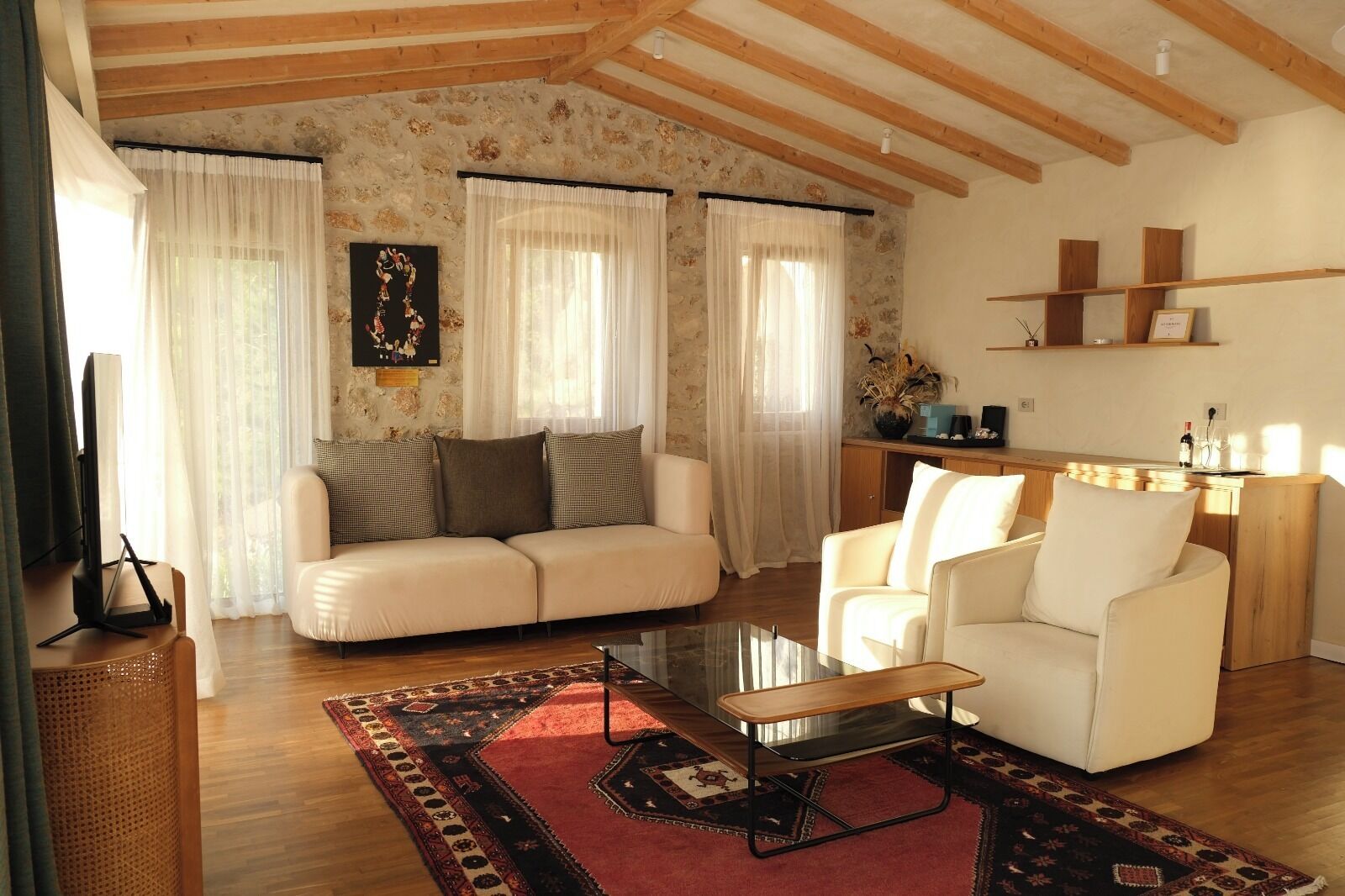 Ein Wohnzimmer mit einer Couch, Sesseln, einem Couchtisch und einem Fernseher. Vorhanden sind Vorhänge, ein Teppich und eine Wand mit Regalen.