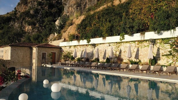 Una piscina con poltrone a sdraio e ombrelloni, un edificio in pietra e una collina sullo sfondo.