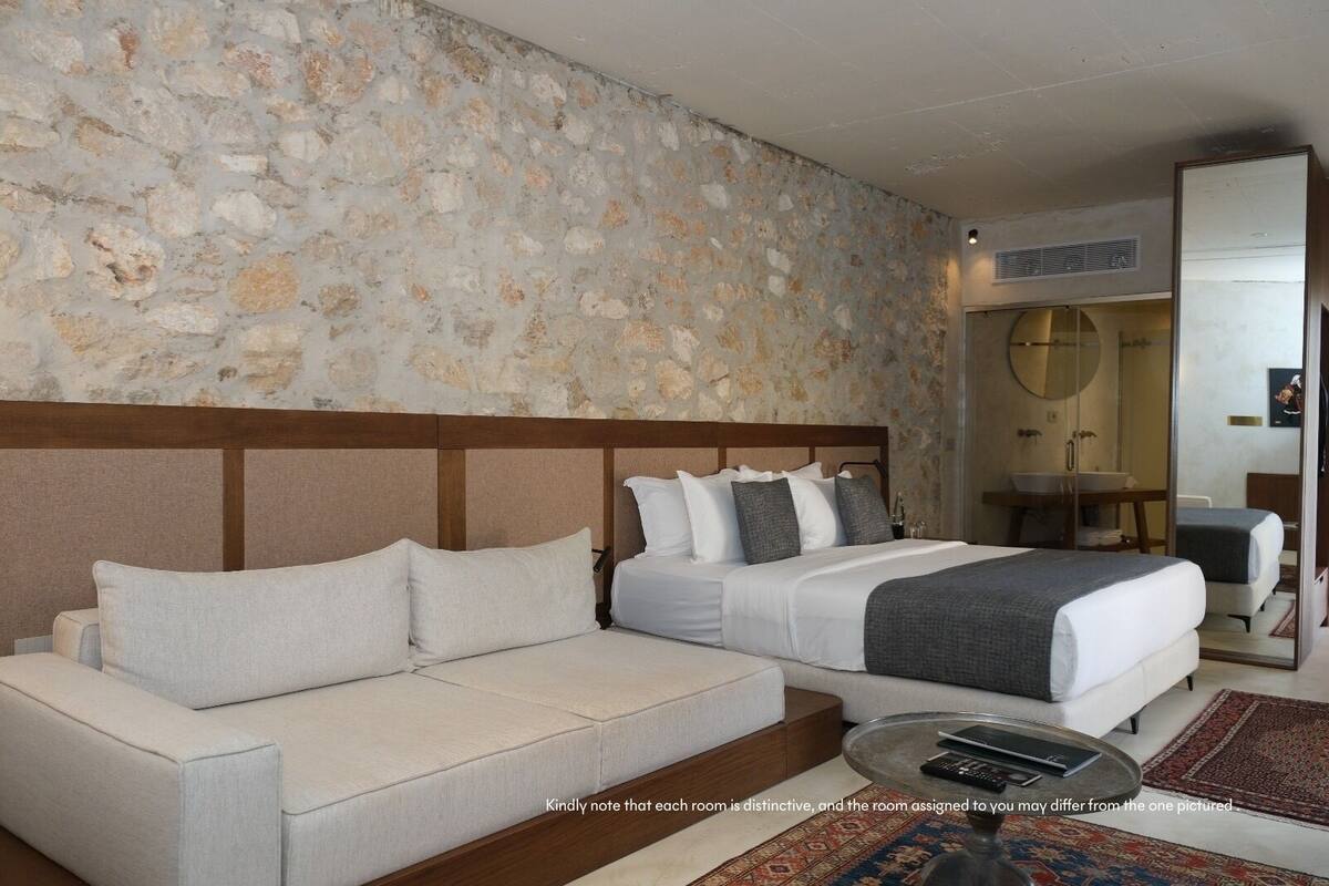 A hotel room with a stone wall, a wooden headboard, a bed with white bedding, a white sofa, a glass coffee table, and a bathroom visible through a glass door.