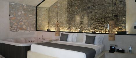 Un dormitorio moderno con pared de piedra, cama grande, bañera e iluminación colgante.