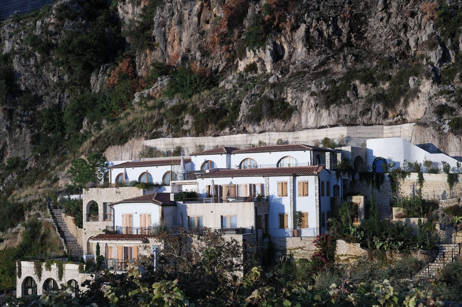 A multi-story building with arched windows and a stone facade, situated on a hillside with a rocky cliff in the background.