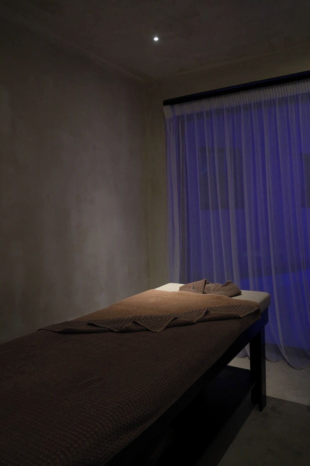 A single bed with a brown blanket in a dimly lit room with a large window covered by blue curtains.