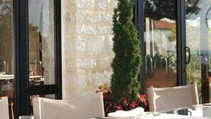 Un ristorante con pareti in pietra e ampie finestre, tavoli all'aperto con tovaglie bianche e sedie in vimini, e un piccolo albero al centro.