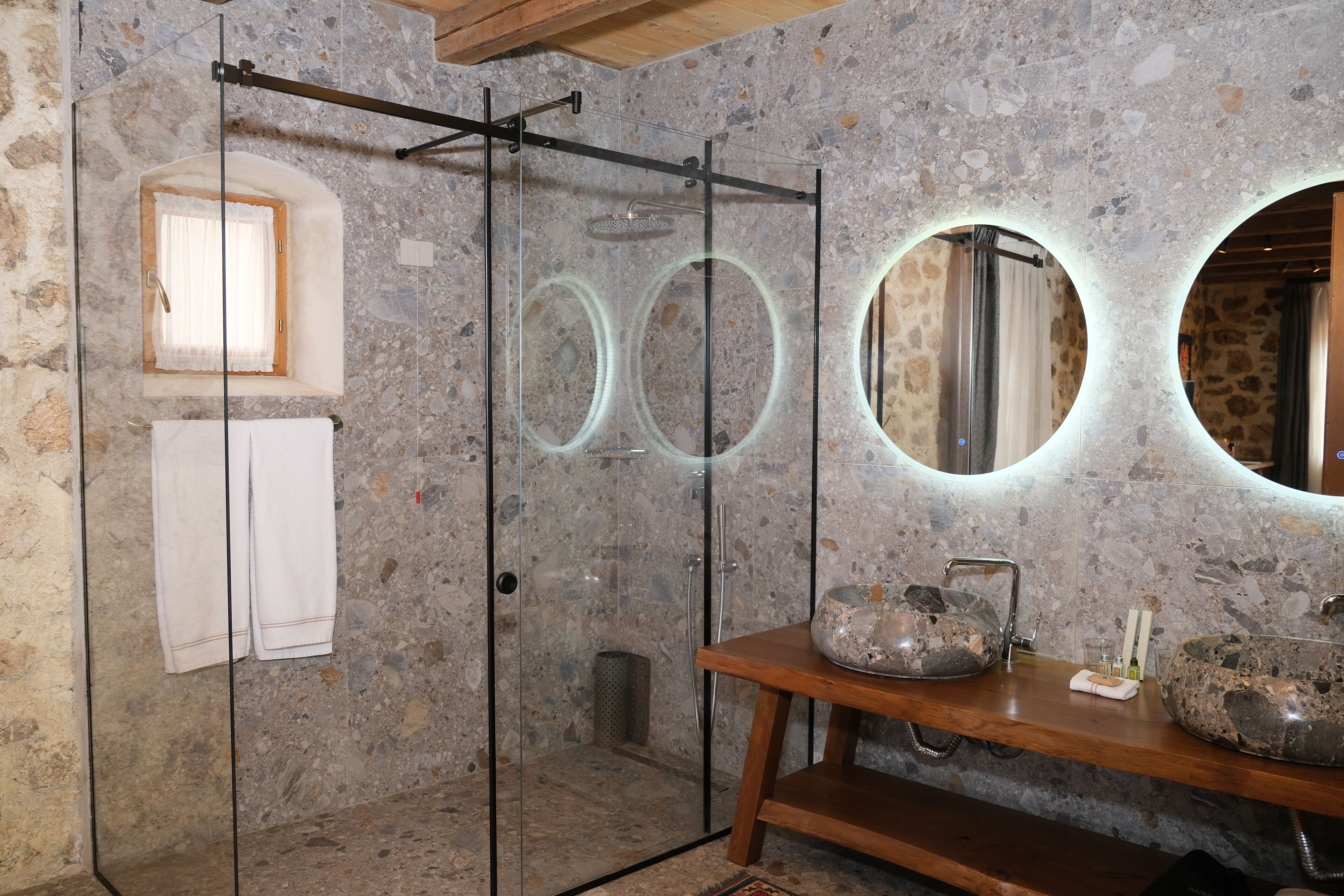 A bathroom with a glass-enclosed shower, stone walls, a wooden vanity with two stone sinks, and a mirror with circular frames.