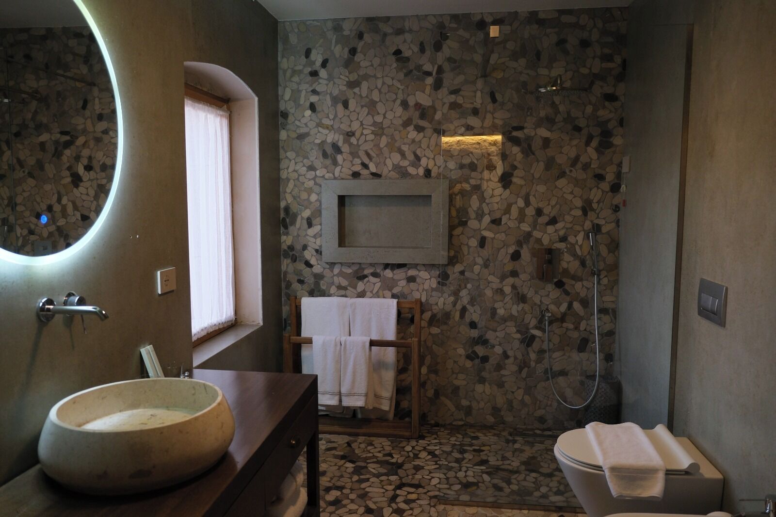 A bathroom with a stone wall, a round mirror, a wooden vanity, a toilet, and a shower area.