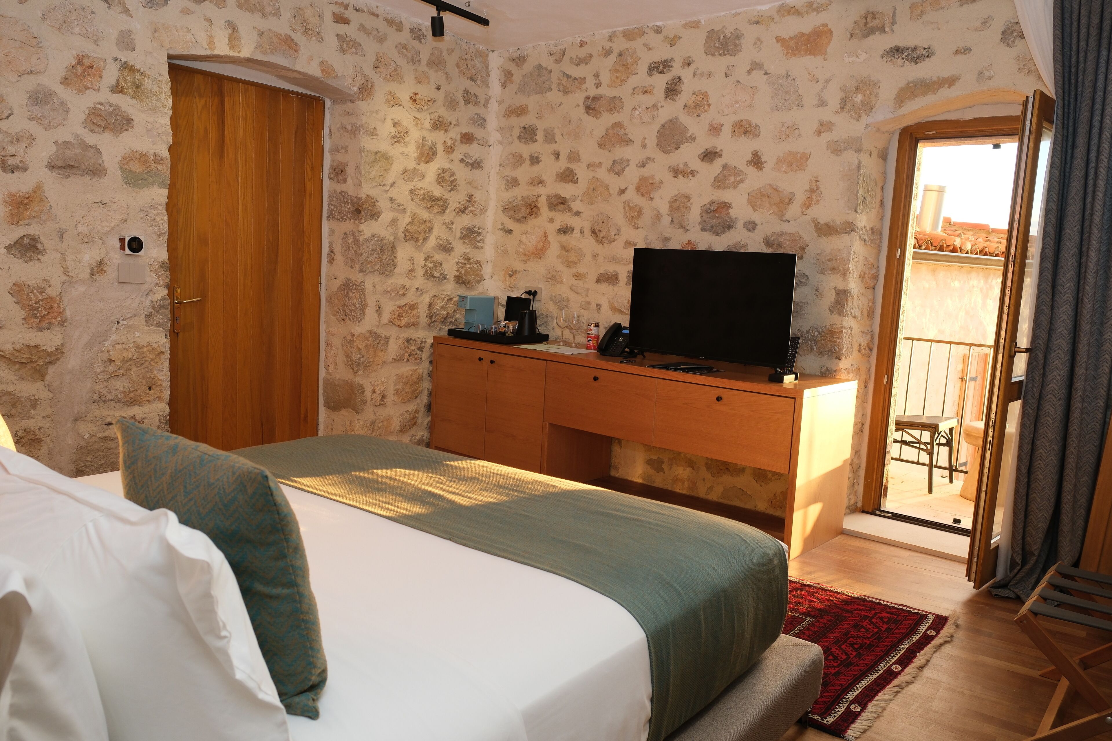 A bedroom with a bed, a wooden desk, a television, and a stone wall.