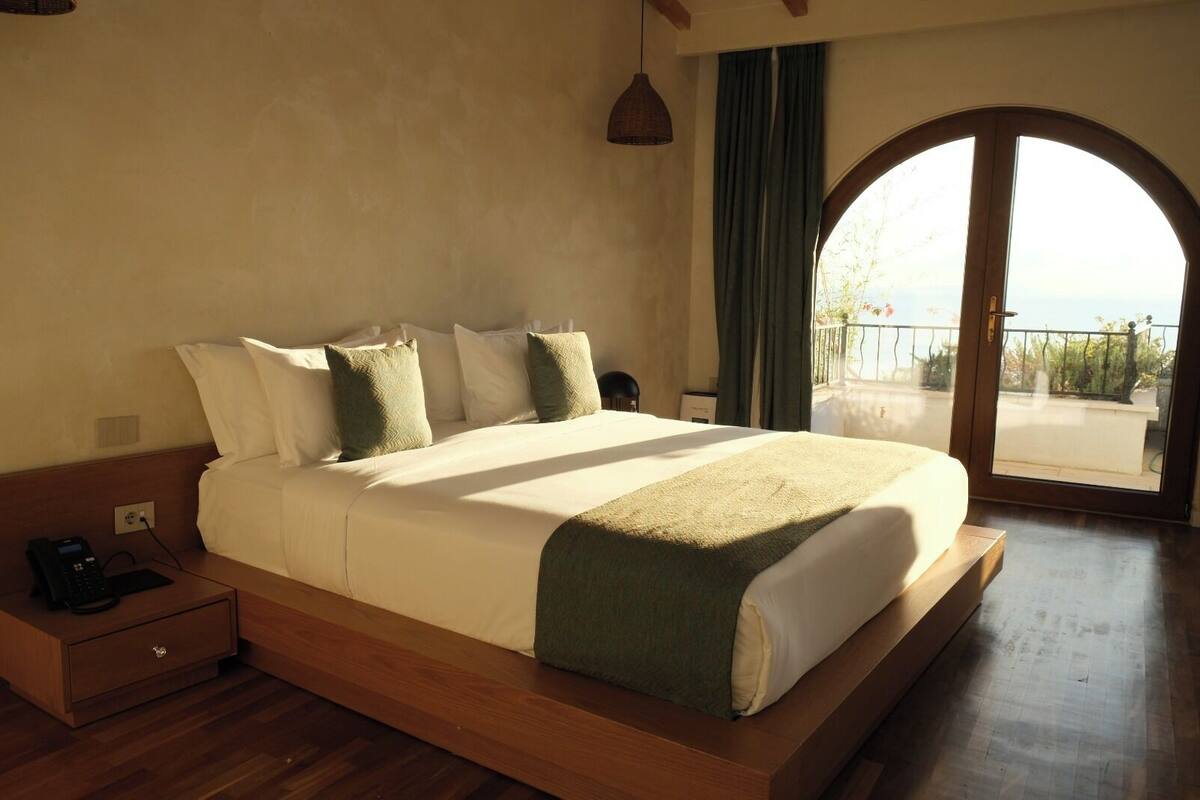 A large bed with a wooden frame, a bedside phone, and a view of a balcony through a large arched window.