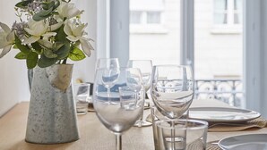 Une table à manger est dressée avec des assiettes, des couverts et des verres à vin, et un vase de fleurs est placé sur le côté gauche.