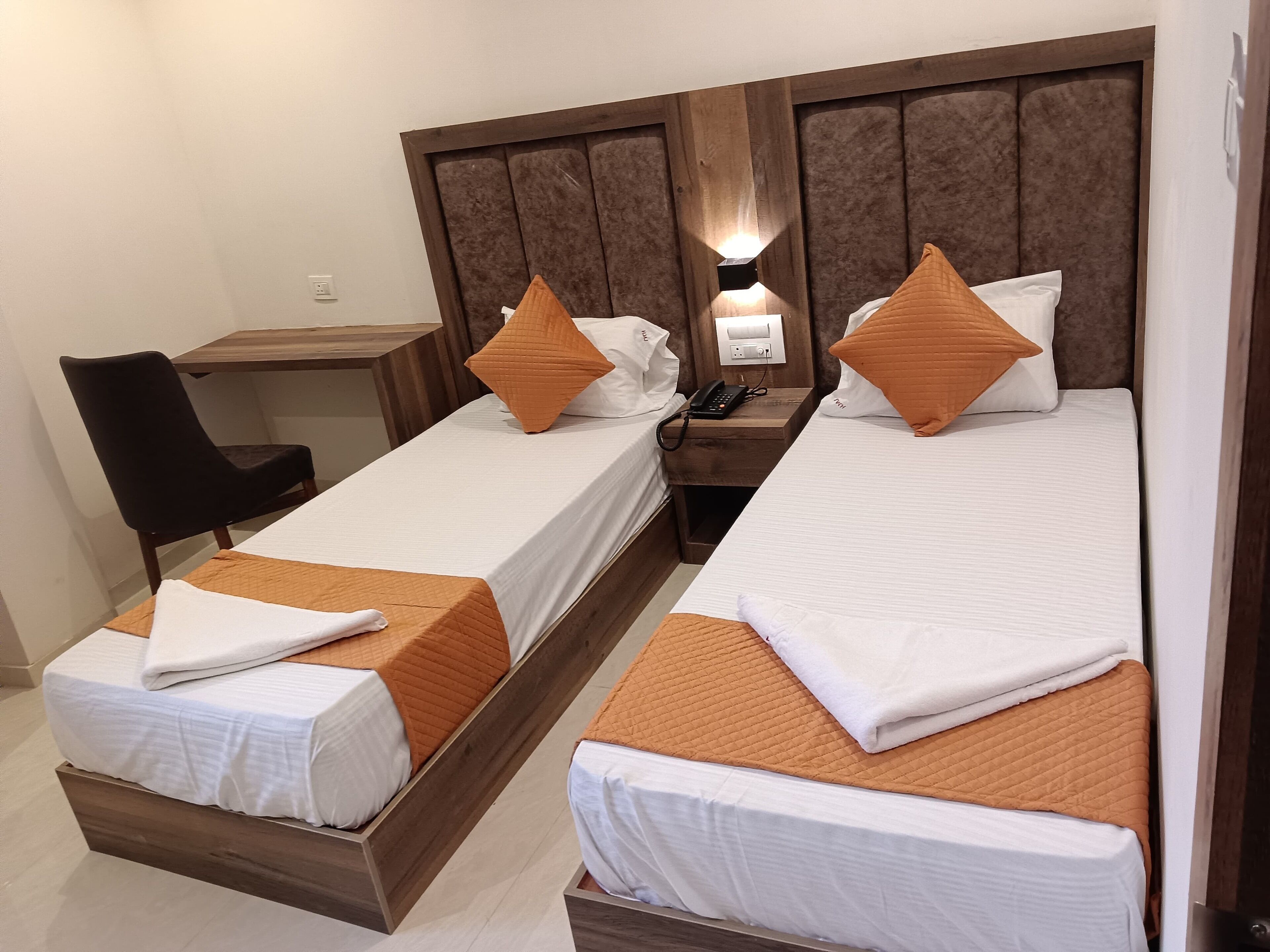 Habitación de hotel con dos camas individuales, cada una con sábanas blancas y colchas anaranjadas, un cabecero de madera, una mesita de noche con un teléfono y una silla.