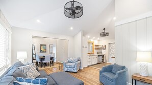 A modern living room with a gray sectional sofa, a patterned armchair, and a blue armchair. There is a dining area with a table and chairs, and a kitchen in the background. - Remodeled, Private Pool & Hot Tub, Beach Nearby (Union Pier)