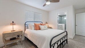 Ein Schlafzimmer mit Bett, Nachttischen, Deckenventilator, Fenster mit Jalousien, Badezimmer mit Spiegel und Waschbecken sowie einer Tür.