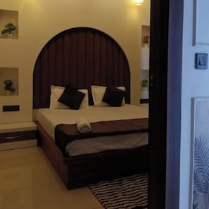 A bedroom with a bed, pillows, a headboard, a nightstand, a wall-mounted shelf with a plant, and a door with a handle. - Hotel Mango Tree (Hansi)