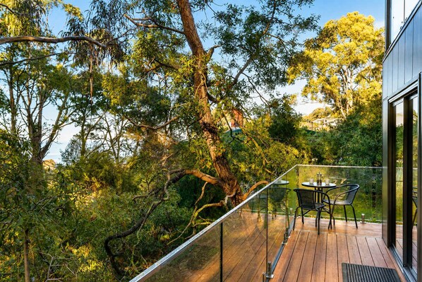 A wooden deck with glass railings overlooking a lush garden with trees and a clear sky.