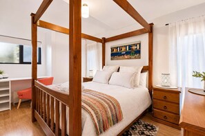 A four-poster bed with a colorful throw blanket, a wooden desk with a chair, a small table with a lamp, and a painting on the wall.