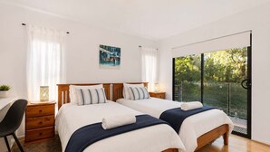 Een hotelkamer met twee bedden, een bureau, een stoel, een kleine tafel met een plant en een groot raam met uitzicht op groen.