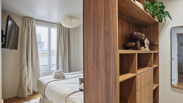 Een slaapkamer met een bed, een raam met gordijnen, een houten plank met decoratieve spullen en uitzicht op buiten.