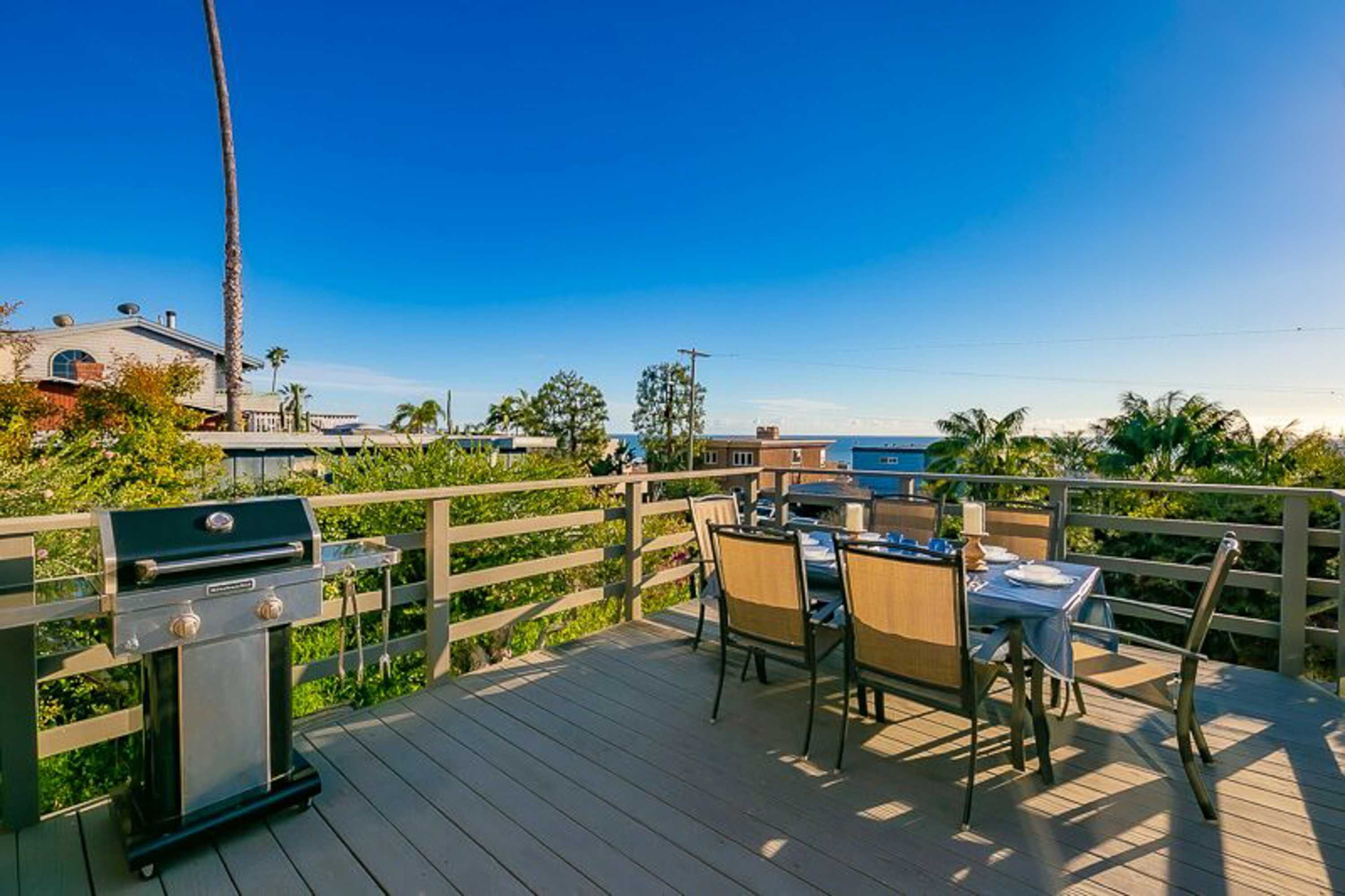 Terraza de madera con una parrilla, sillas y una mesa, con vistas a un cuerpo de agua y un edificio.