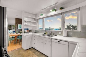 A modern kitchen with white cabinetry, a built-in dishwasher, and a window with a view of the sea. - Ocean View Deck! | Walk to the Beach | King Suite (Encinitas)