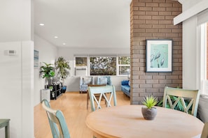 A modern living room with a round wooden table, two chairs, a sofa, and a large window with a view of trees. - Ocean View Deck! | Walk to the Beach | King Suite (Encinitas)