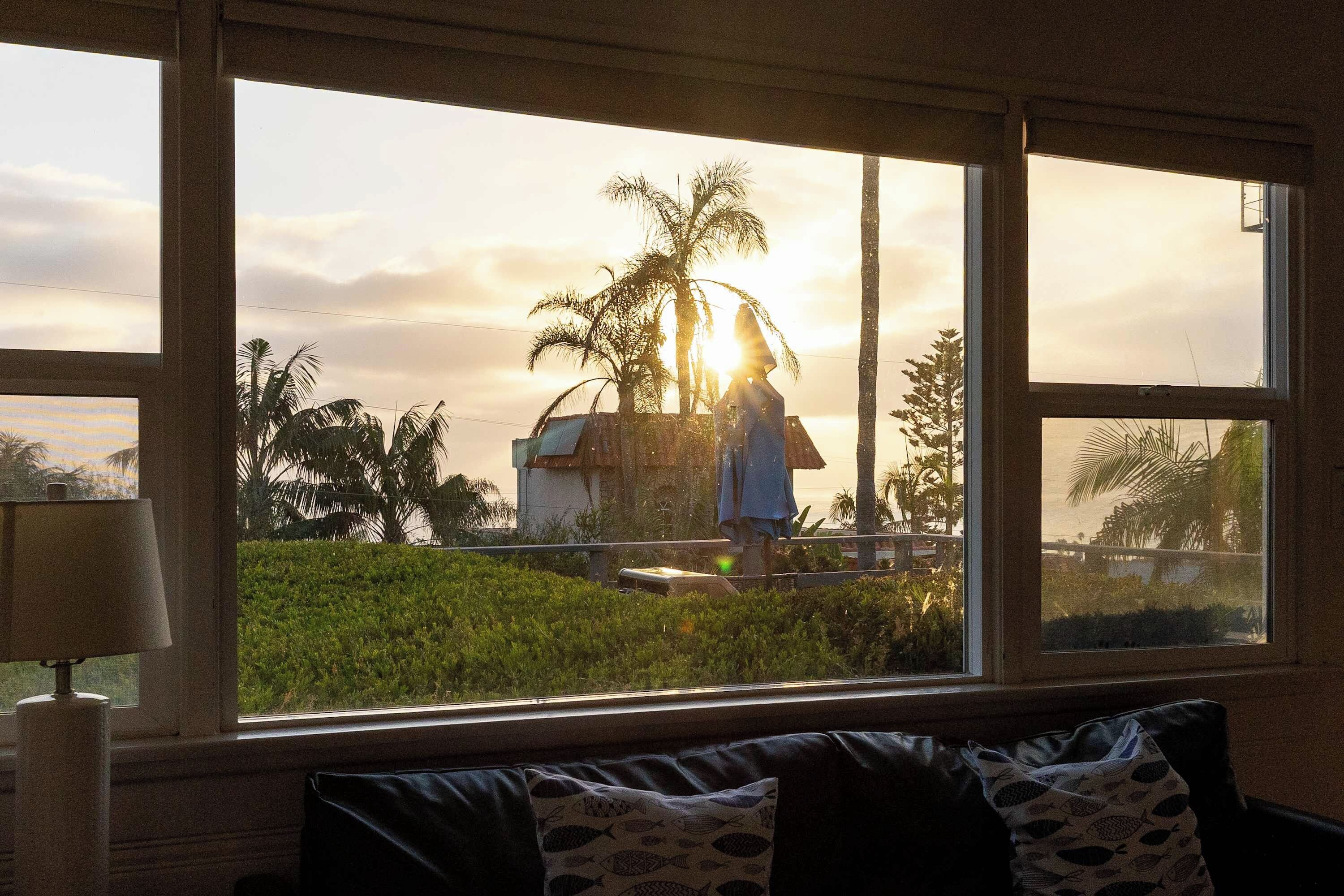 A room with a large window overlooking a landscape with palm trees and a building, a lamp on a table, and a sofa with patterned cushions.