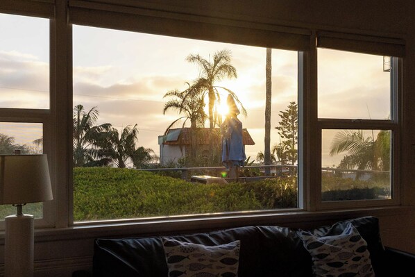 A room with a large window overlooking a landscape with palm trees and a building, a lamp on a table, and a sofa with patterned cushions. - Ocean View Deck! | Walk to the Beach | King Suite (Encinitas)