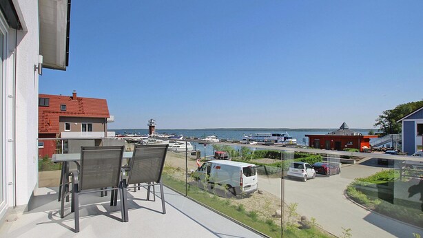 Ein Balkon mit Tisch und Stühlen mit Blick auf einen Yachthafen, wo Boote und Autos geparkt sind.