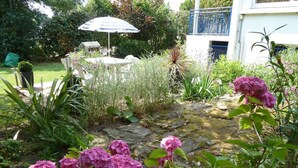 Un jardín con un camino de piedra, hortensias rosas y una mesa blanca con un paraguas a rayas.