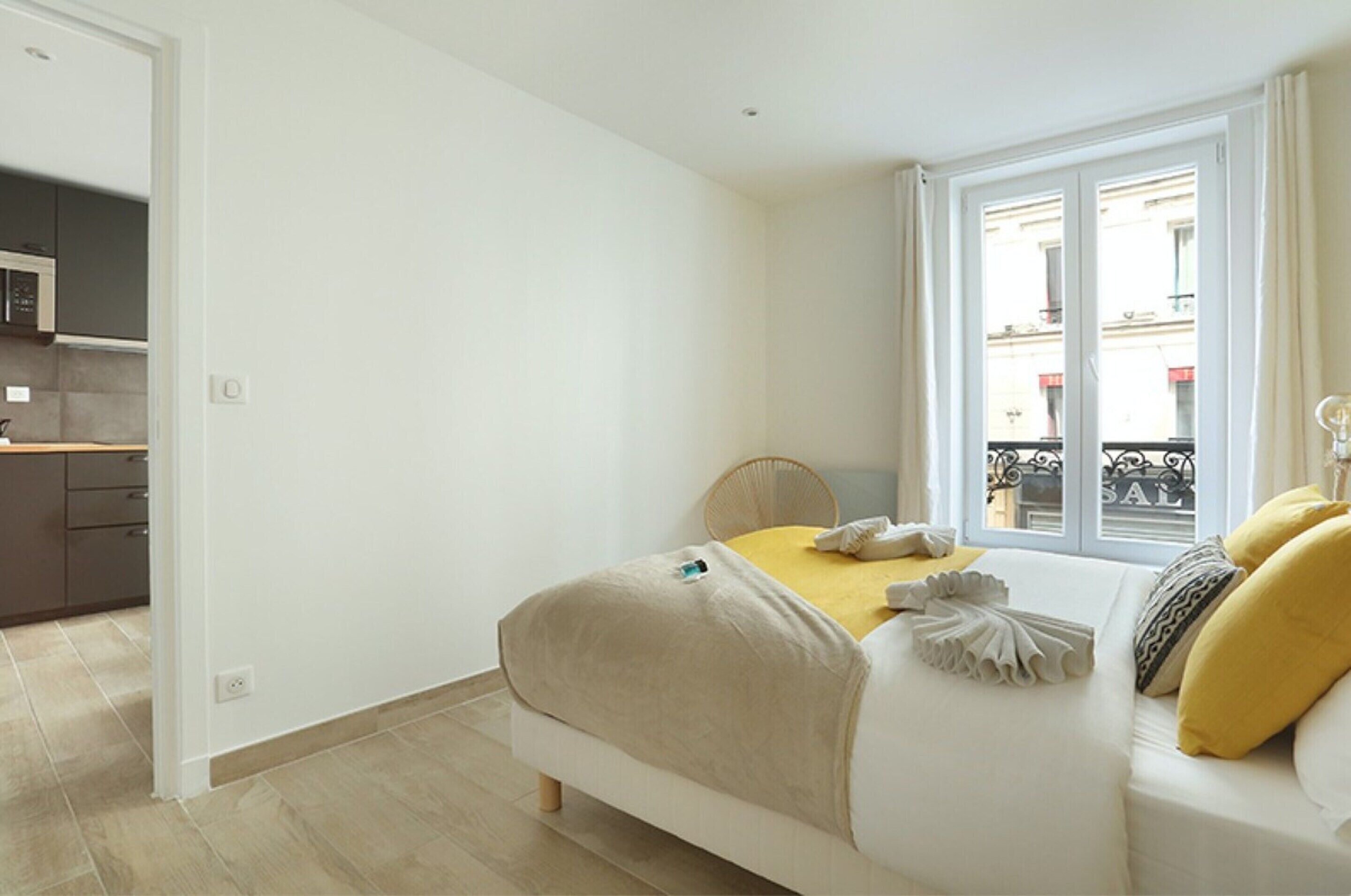 A bedroom with a bed, a window with a view of buildings, and a kitchenette in the background.