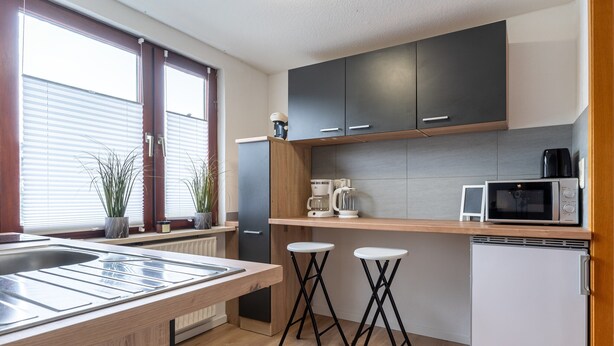 A modern kitchen with a wooden countertop, a built-in microwave, and a refrigerator. There are two stools and a window with blinds. - Ferienwohnung in Busum (Büsum)