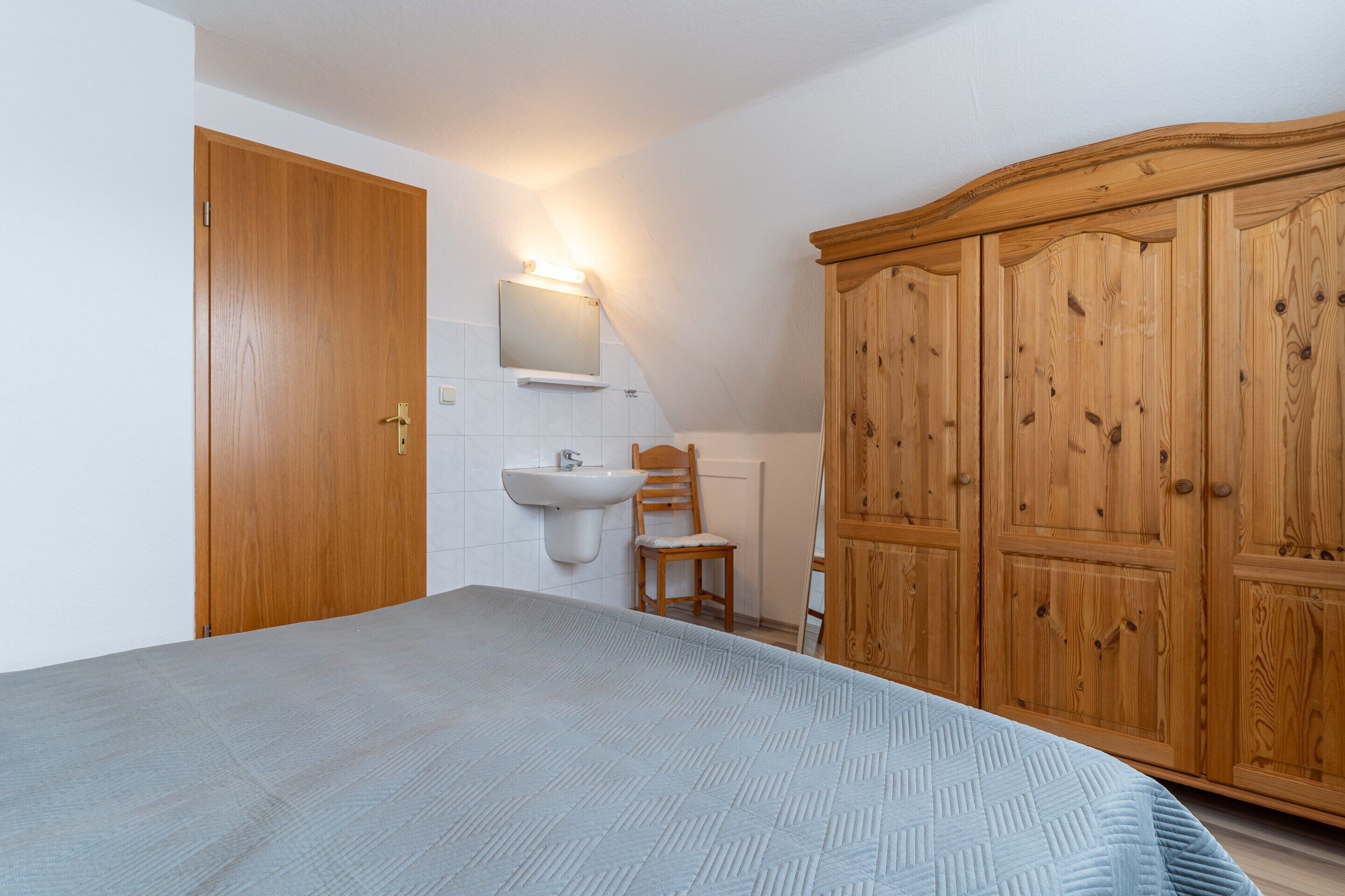 A bedroom with a bed, a wooden wardrobe, a wooden chair, a white sink, and a mirror.