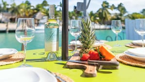 Mesa posta para refeição com garrafa de vinho, pratos, talheres e frutas tropicais.