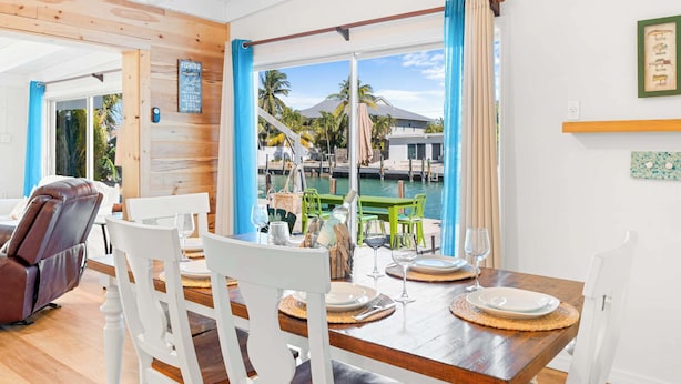 Uma sala de jantar com mesa de madeira posta para quatro, cadeiras brancas e vista para uma marina com barcos e palmeiras.