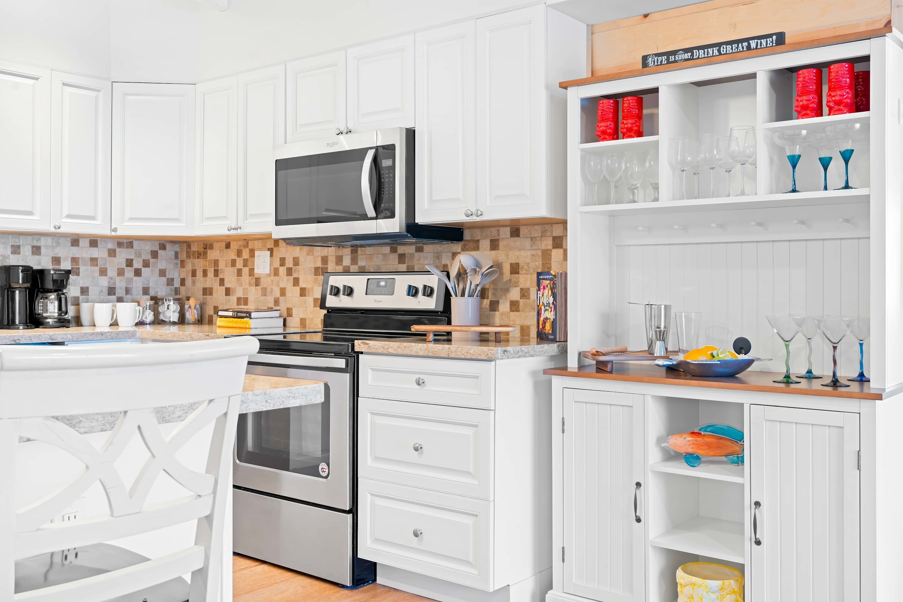 Cocina moderna con armarios blancos, microondas, horno y cafetera. Hay una repisa con copas de vino y un cartel que dice 'IT'S SO GOOD, DRINK GREAT WINE!'.