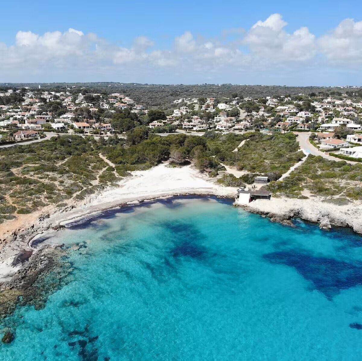 An aerial view of a coastal area with a beach, clear turquoise waters, and a residential area with houses.