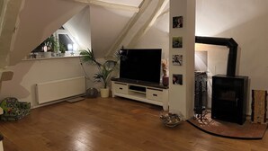 A cozy attic room with a wooden floor, a large flat-screen TV, a fireplace, and a window with a plant. - Vacation apartment  (Vettweiß)
