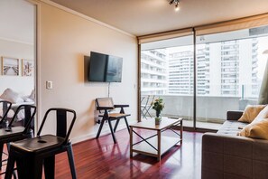 A modern living room with a flat-screen TV, a dining table, a sofa, and a balcony with a city view. - Comfortable 1BR Apt. in Prime Las Condes Location (Las Condes)